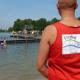 Bademeister von hinten mit organgenem Hemd und Freibad-Aufdruck vor dem See